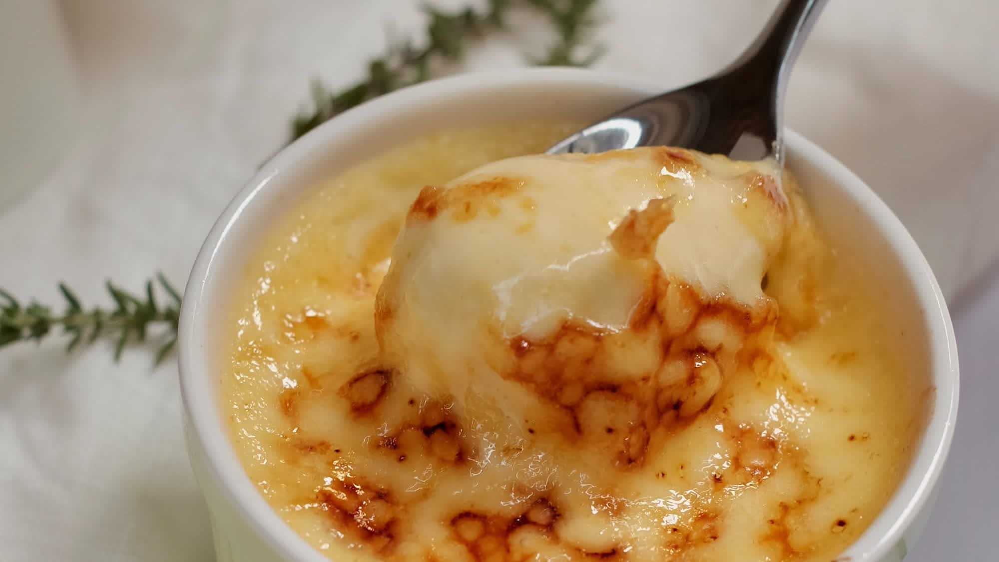 Crème brulée in einem weißen Förmchen mit goldbraun gebräunter Kruste, ein Löffel hebt eine Portion heraus, daneben Thymianzweige auf einem weißen Tuch