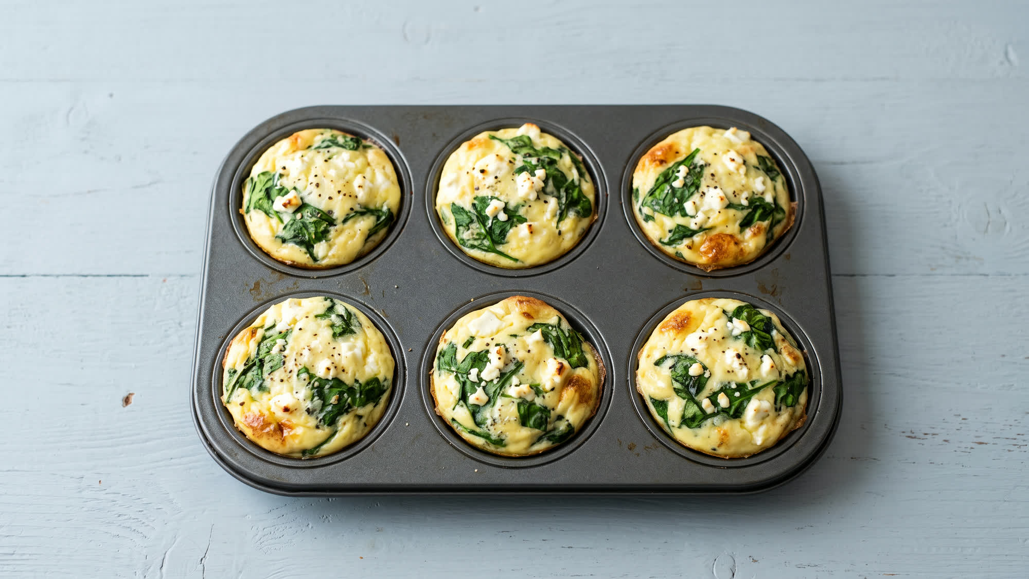 Sechs hellgelbe Ei-Muffins mit Spinat und zerbröseltem Feta in einem dunklen Muffinblech auf einem hellblaugrauen Holztisch