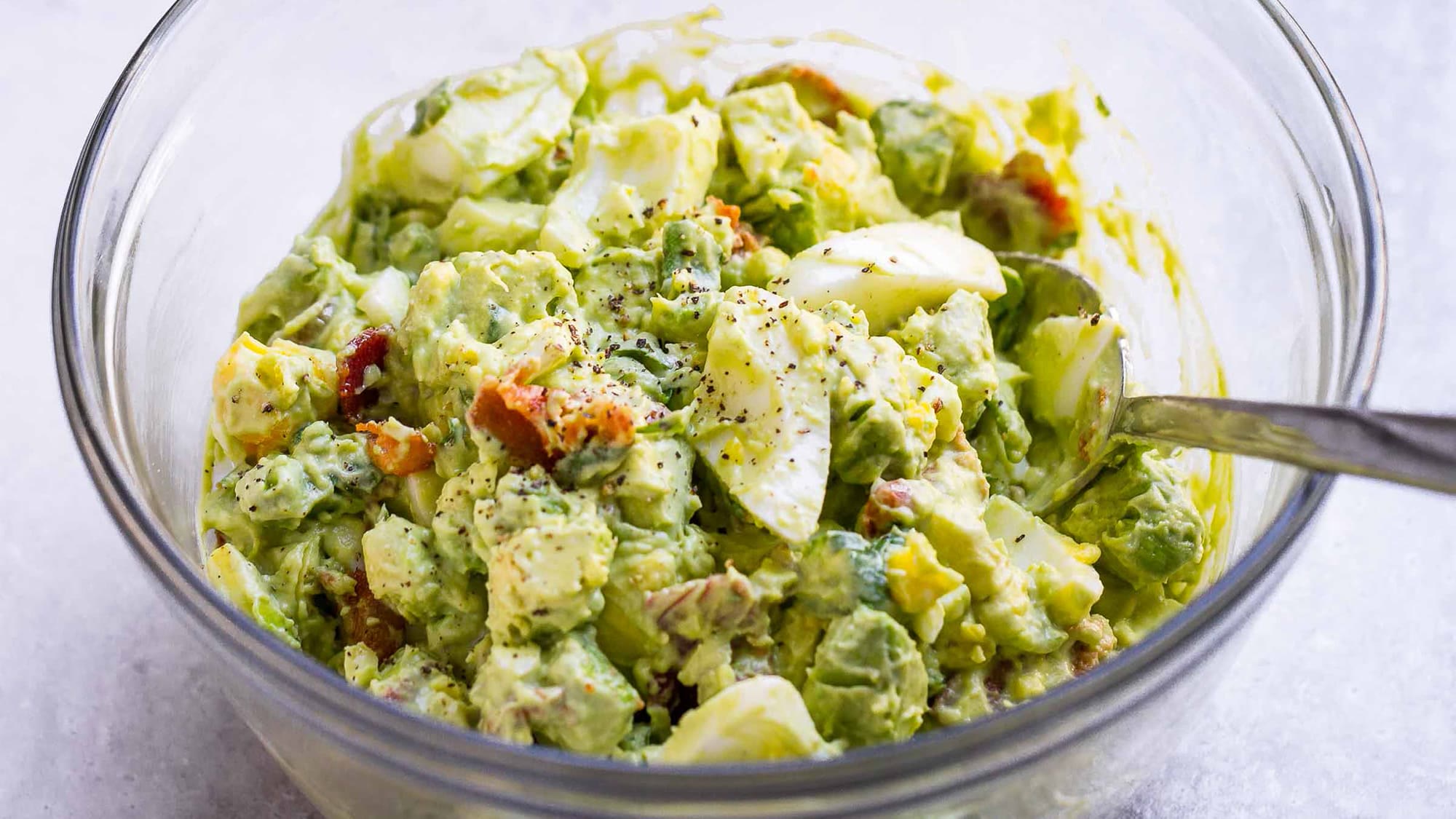 Eiersalat mit Avocado in einer Glasschüssel mit Löffel, darin auch Speckstücke, Kräuter und schwarzer Pfeffer