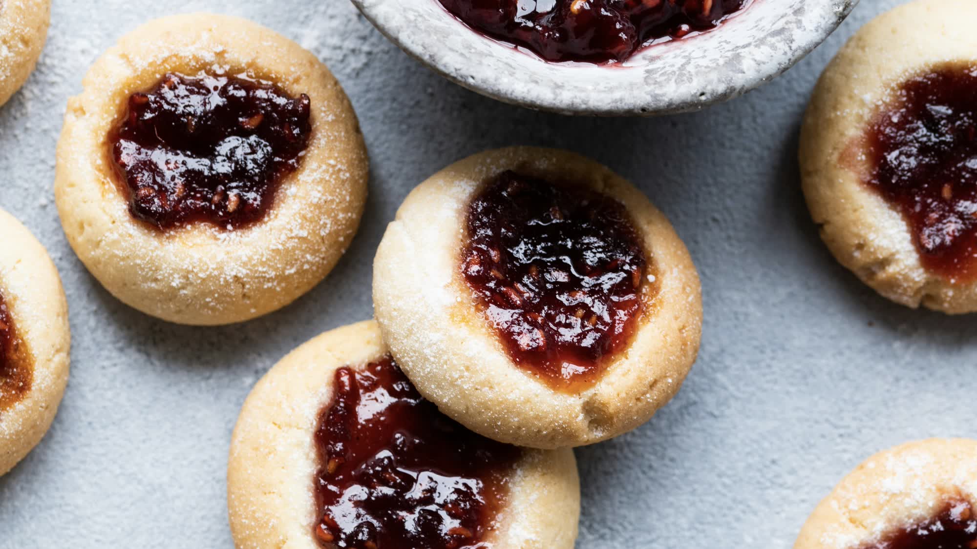 Runde, helle Engelsaugen mit dunkelroter Himbeermarmelade in der Mulde, mit Puder bestäubt, auf hellgrauem Untergrund, oben eine angeschnittene Schale mit Marmelade