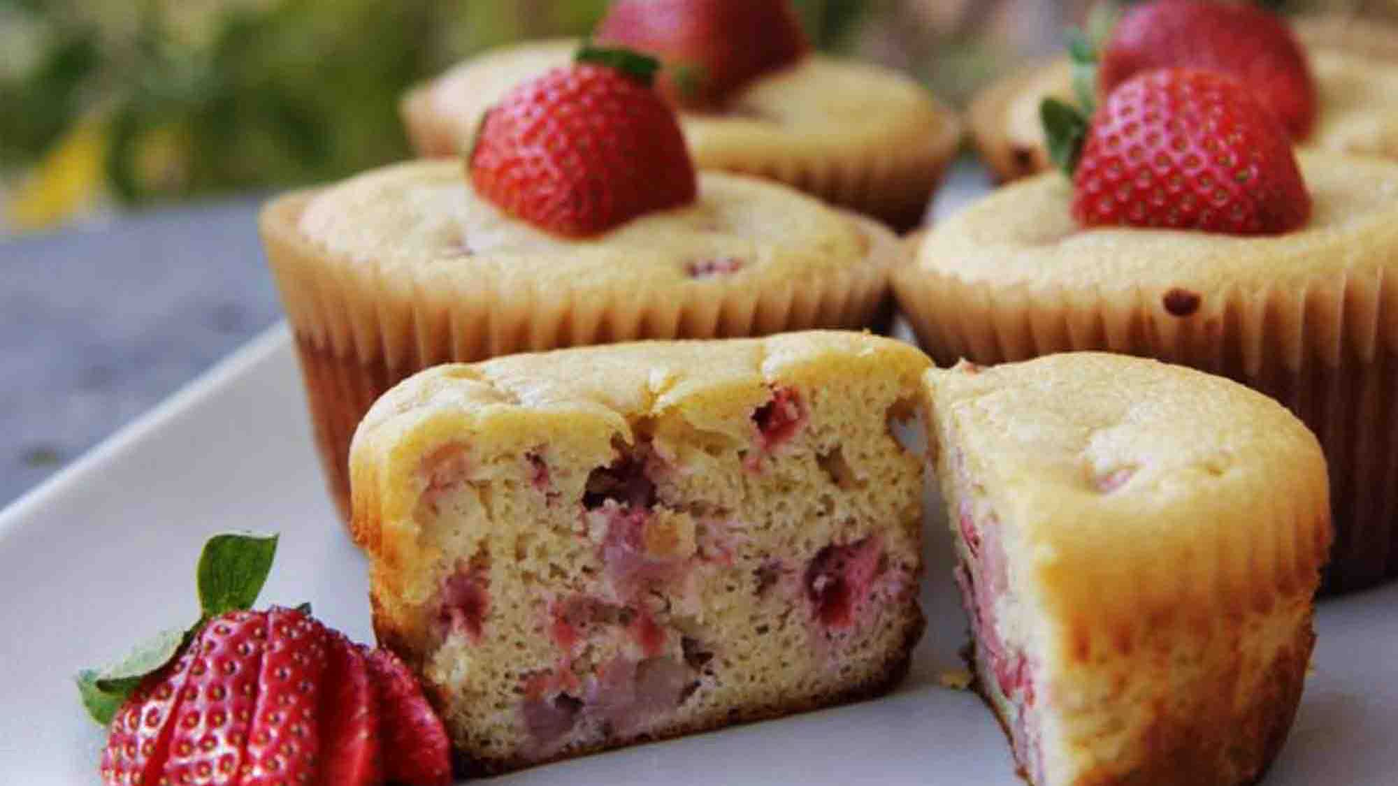 Goldgelbe Muffins in hellen Papierförmchen mit Erdbeerstücken im Teig, obenauf je eine ganze Erdbeere, vorn ein aufgeschnittener Muffin und eine aufgefächerte Erdbeere