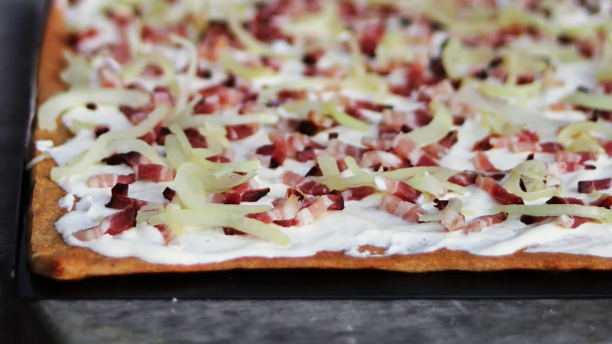 Flammkuchen auf einem Backblech: goldbrauner Boden mit Crème fraîche, Speckwürfeln und hellen Zwiebelstreifen