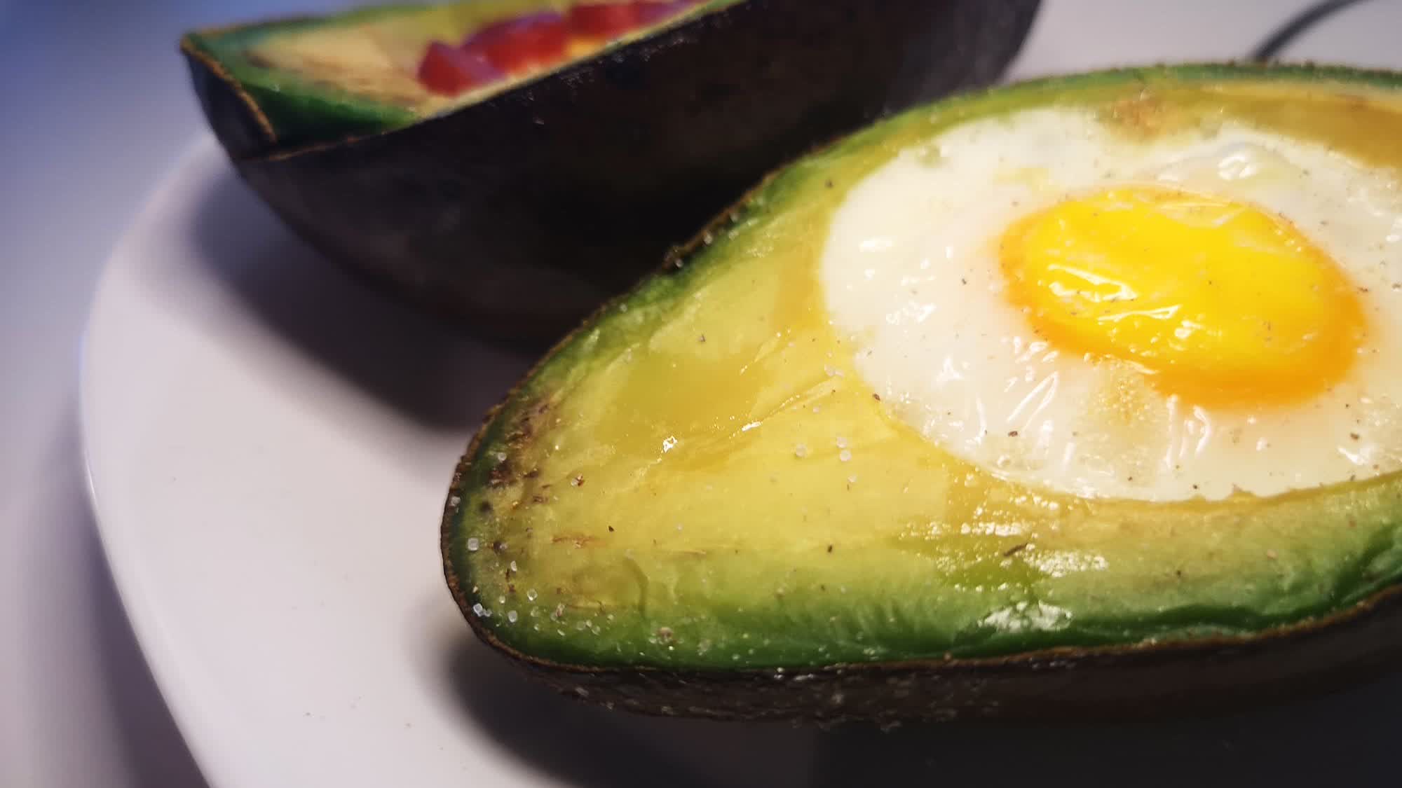 Gebackene Avocadohälfte auf einem weißen Teller mit einem ganzen Ei in der Mulde, mit Salz und Pfeffer bestreut, dahinter eine zweite Avocadohälfte mit roten Würfeln