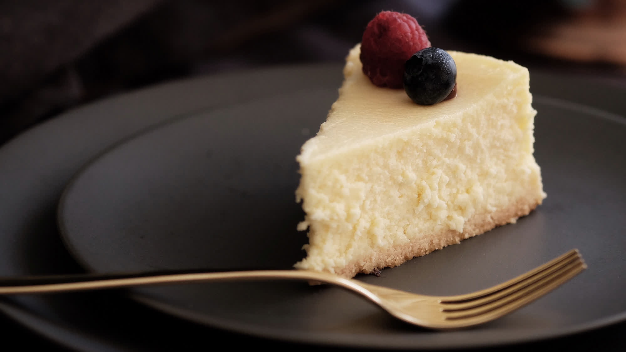 Ein Stück gebackener Käsekuchen mit heller Füllung und dünnem, hellem Boden auf einem dunklen Teller, obenauf eine Himbeere und eine Heidelbeere, davor eine goldene Gabel