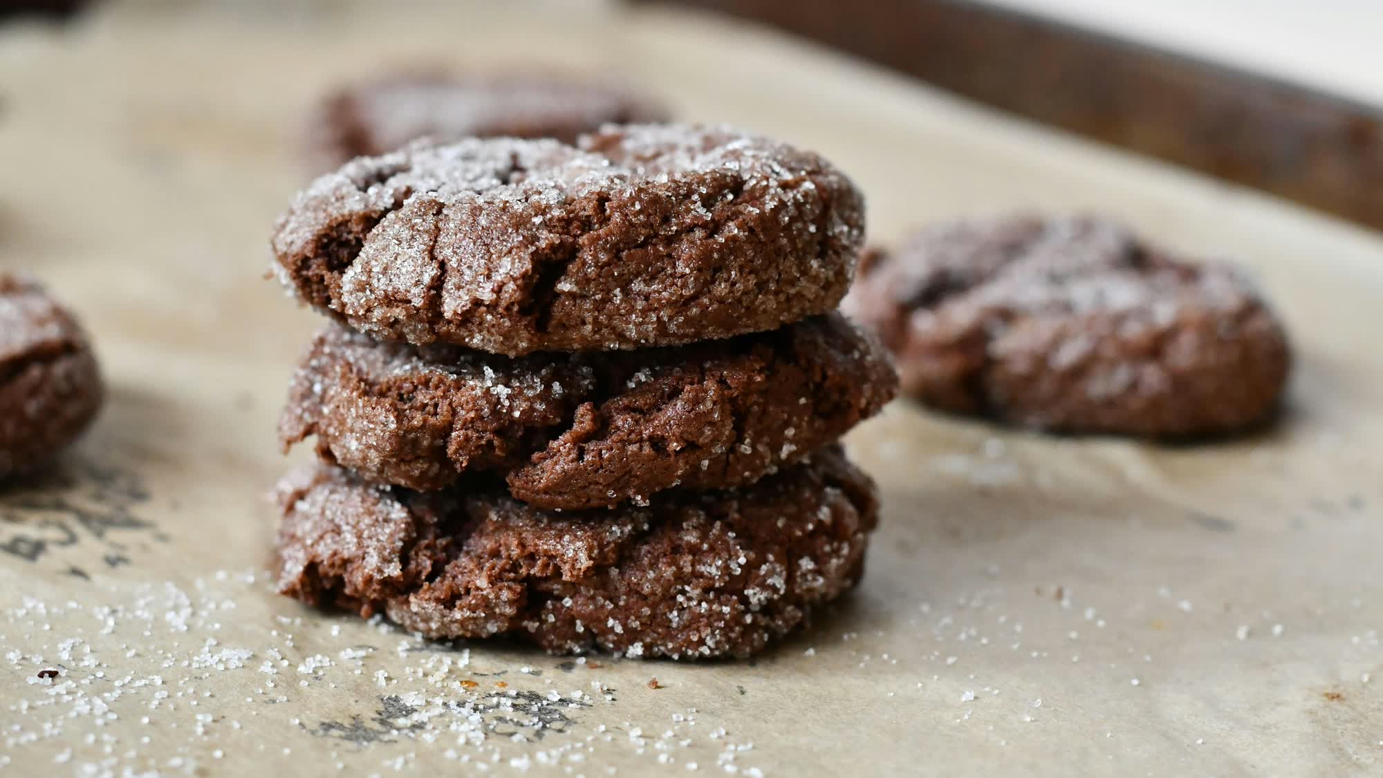 Drei gestapelte Schokocookies mit weißen Kristallen bestreut auf Backpapier, dahinter weitere Cookies