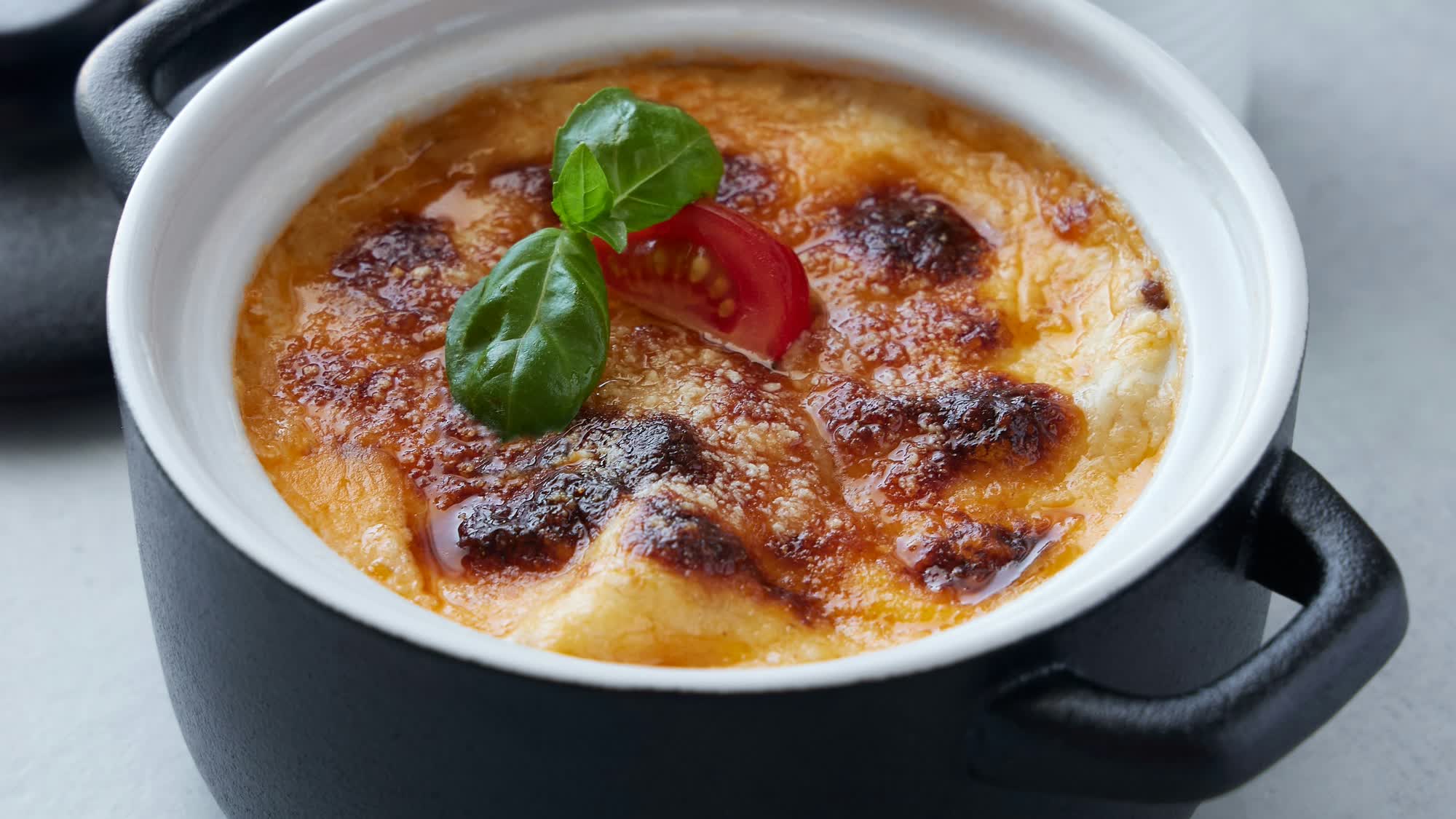 Überbackener Auflauf mit goldbrauner Käsekruste in einem schwarzen Töpfchen mit weißem Rand, obenauf eine Tomatenspalte und Basilikumblätter, auf hellem Untergrund