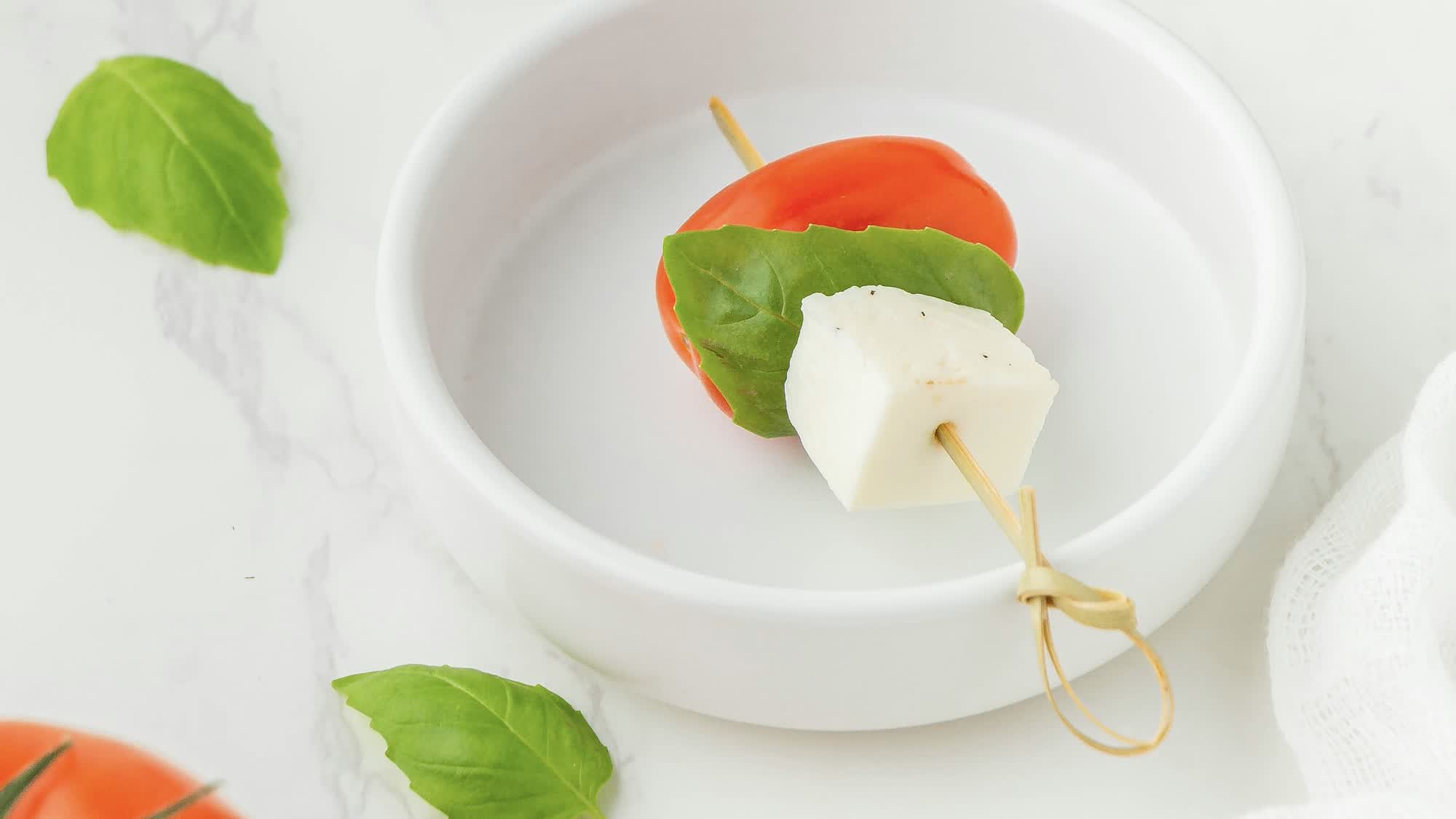 Spieß mit Cherrytomate, Basilikumblatt und Mozzarella-Würfel in einem weißen Schälchen
