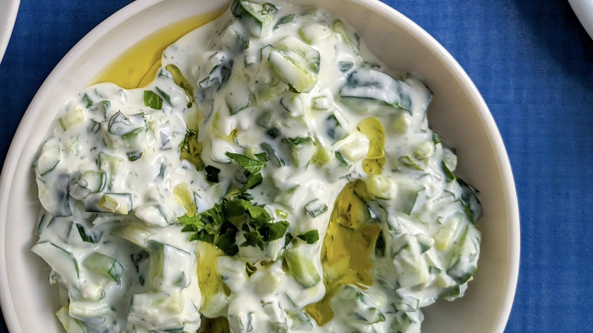 Tzatziki in einer weißen Schale mit Gurkenstücken, Olivenöl und gehackter Petersilie