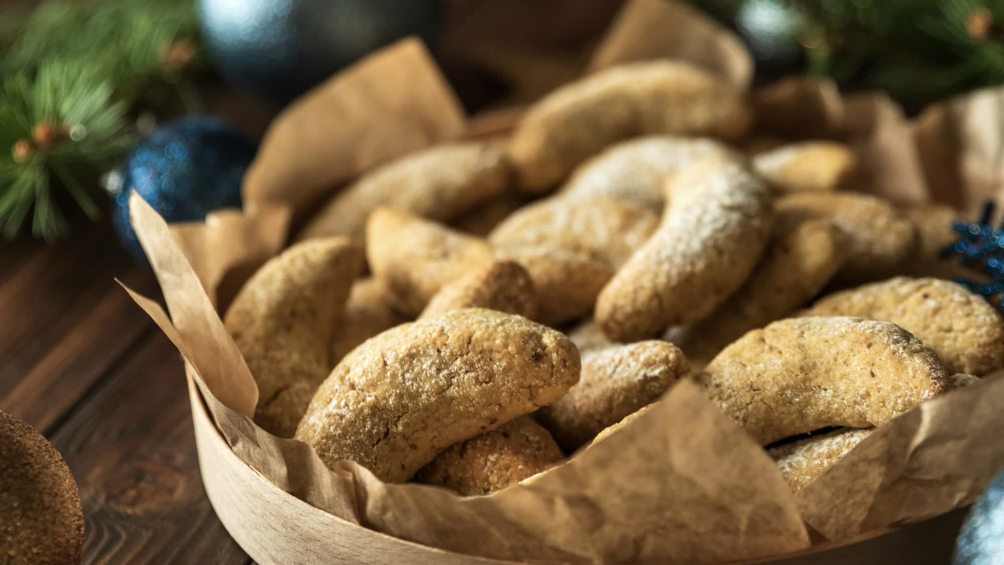 Vanillekipferl mit hellem Puder in einer Holzschachtel mit Backpapier, daneben Christbaumkugeln und Tannenzweige