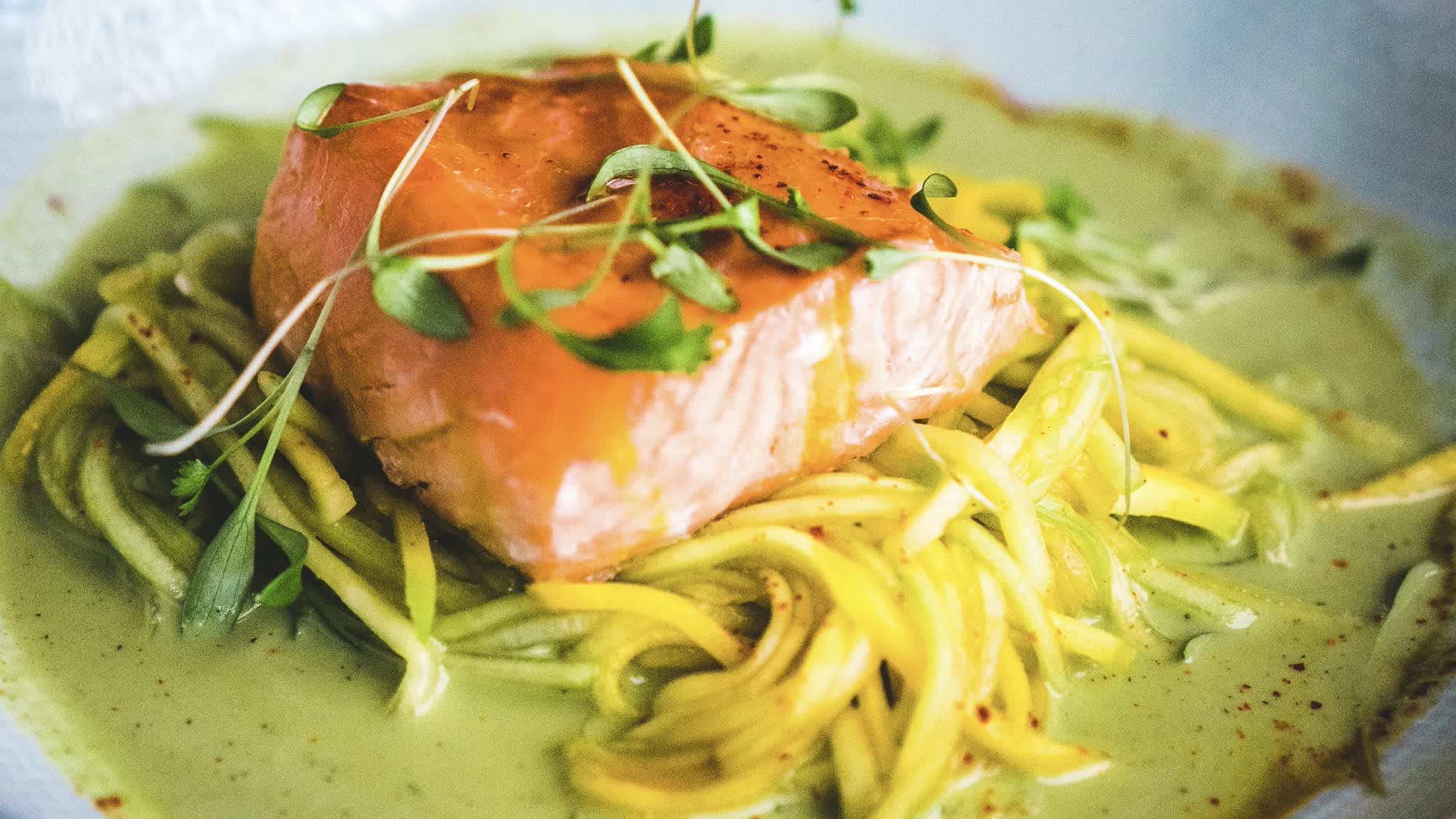 Ganzes Lachsfilet auf Zucchininudeln in reichlich grüner Soße mit Microgreens und roten Gewürzflocken in einer weißen Schüssel