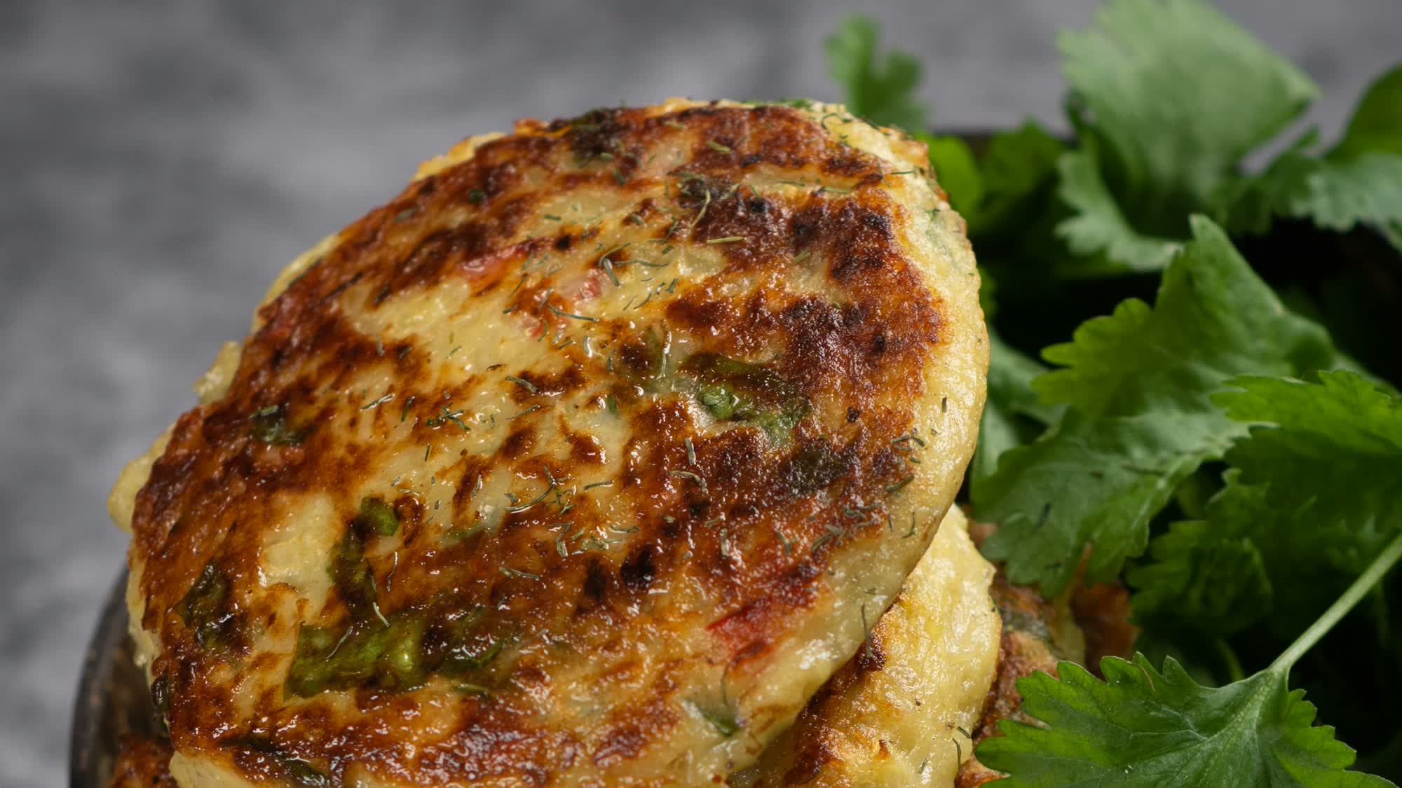 Gestapelte goldbraune Zucchini Puffer mit Kräutern und kleinen roten Stücken im Teig, mit Dill bestreut, in einer dunklen Schale mit frischen Korianderblättern