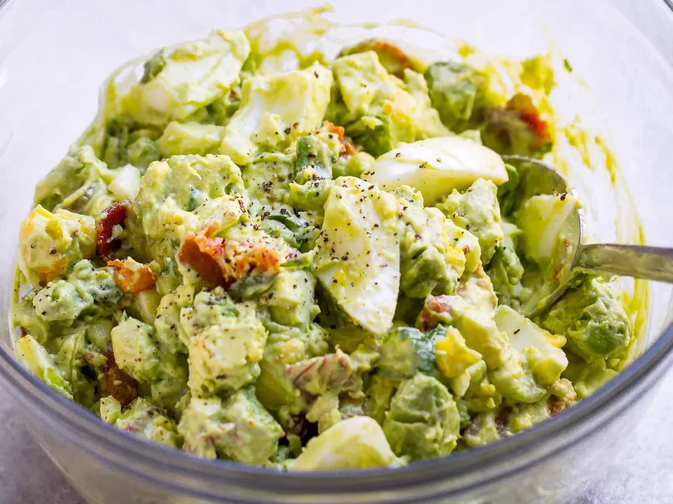 Eiersalat mit Avocado in einer Glasschüssel mit Löffel, darin auch Speckstücke, Kräuter und schwarzer Pfeffer