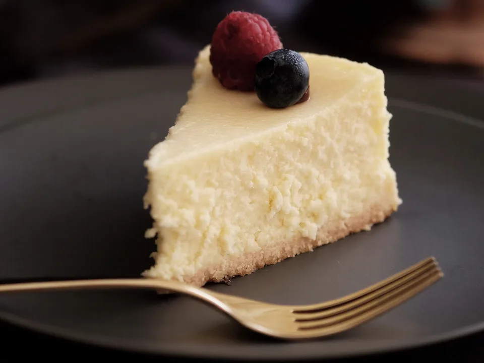 Ein Stück gebackener Käsekuchen mit heller Füllung und dünnem, hellem Boden auf einem dunklen Teller, obenauf eine Himbeere und eine Heidelbeere, davor eine goldene Gabel