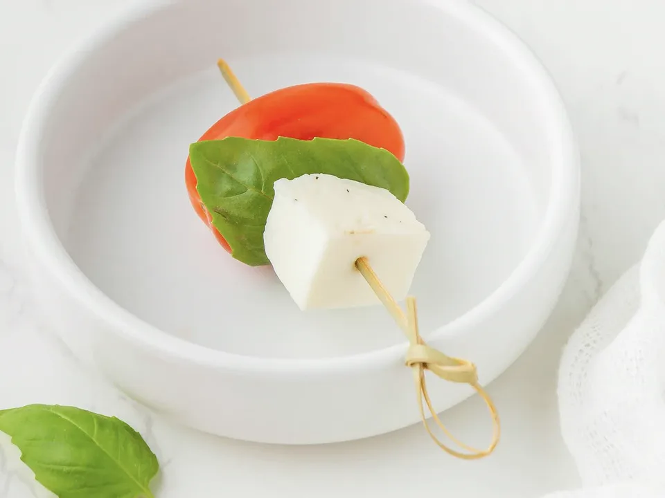 Spieß mit Cherrytomate, Basilikumblatt und Mozzarella-Würfel in einem weißen Schälchen