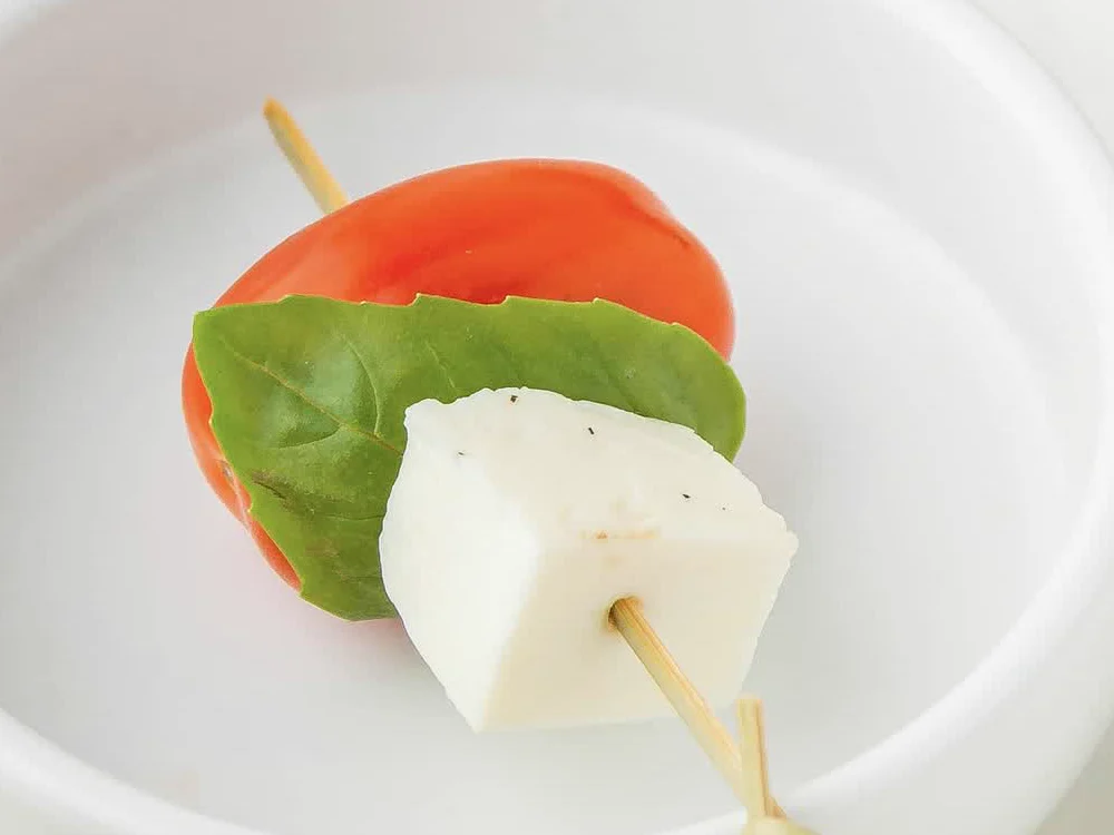 Nahaufnahme eines Spießes mit Kirschtomate, Basilikumblatt und Mozzarella-Würfel in einer weißen Schale
