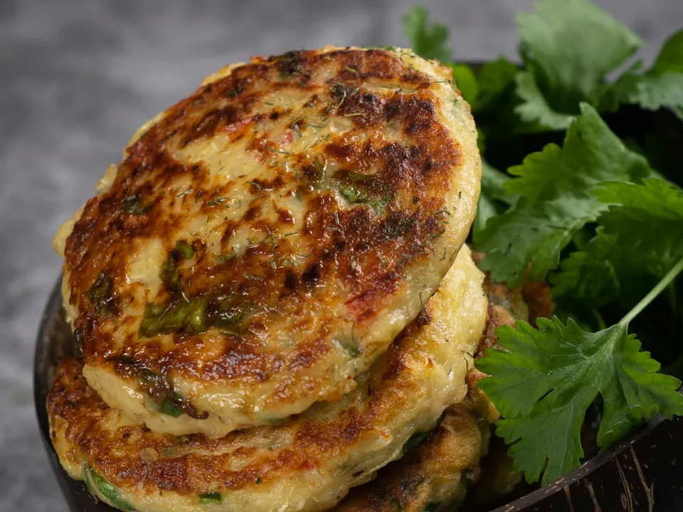 Gestapelte goldbraune Zucchini Puffer mit Kräutern und kleinen roten Stücken im Teig, mit Dill bestreut, in einer dunklen Schale mit frischen Korianderblättern